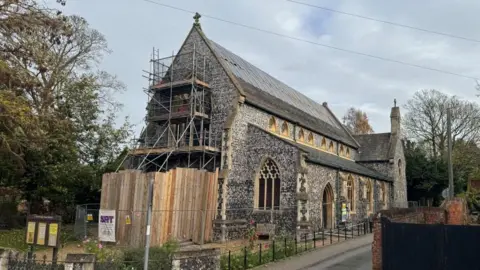 Andrew Turner/BBC A Victorian church built of square knapped flint, with arched windows with sandstone mullions and a slate roof. The building has been damaged by fire, with plastic sheeting and wooden battens holding temporary repairs in place on the roof. Fencing can be seen around the building, with trees behind.