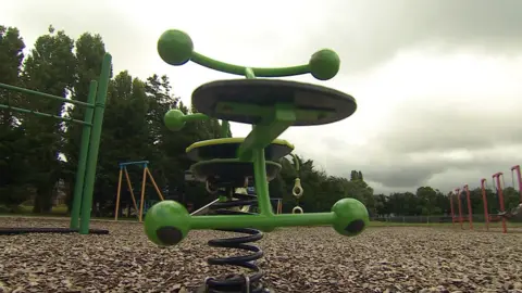 A piece of play park equipment - a spring rider - it is bright green and has two seats. The rest of the park can be seen in the background, with dark clouds overhead. 