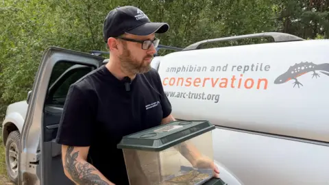 A man holding a plastic box with a lizard inside it. In the background there is a van with the words "amphibian and reptile conservation" on the side.