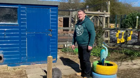 Rick, who has a crew cut, is stood in a rabbit enclosure and is wearing a green hoodie. There is a blue shed next to him and it is sunny.
