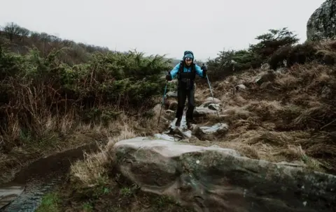 Clorroe Cam/Montane Spine Race A woman with poles runs along a rocky surface