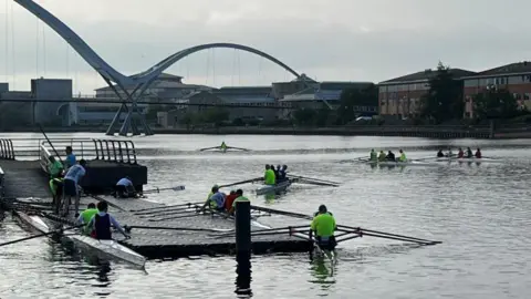 Seven rowing boats on the water which are full of rowers wearing fluorescent clothing. The River Tees is wide and is silver-coloured in the light. Some boats are travelling down the river, while other rowers are preparing to set off. 