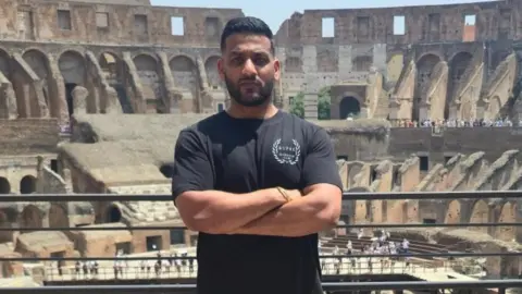 Bally Cheema A man is wearing a black T-shirt and is standing in front of the colosseum in Rome. He has a short dark beard and short dark hair.