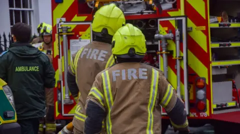 Fire crews attend a scene alongside paramedics. Two firefighters are seen with brown and yellow jackets with "FIRE" written in capital letters on the back and luminous yellow helmets