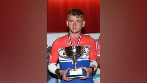 360cycling Aidan Worden - a  young man with short, blondish hair and a medal around his neck, holds a trophy. He wears a red, white and blue cycling jersey. 