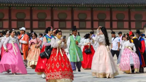 Getty Images Dozens of tourists wearing traditional Korean clothing - called hanbok - visit Gyeongbokgung Palace in Seoul. A section of Gyeongbokgung Palace - a row of doors - can be seen behind groups of people wearing colorful clothing.