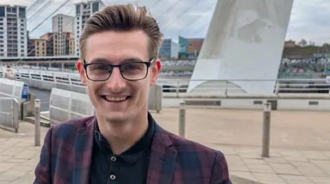 Rob Reed smiles at the camera with Newcastle quayside in the background. He's wearing a checked blazer with blues and purples and his hair is styled in a neat quiff. He has black-rimmed glasses.