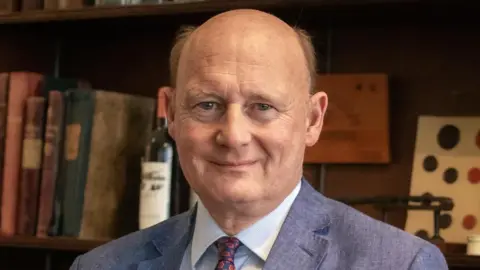 James Tanner James Tanner wearing a blue suit with a floral tie. There is a bottle of wine and books on a shelf behind him.