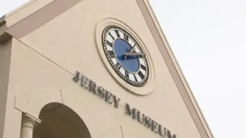 A close up of the front of Jersey Museum. There is a large clock on a light brown building with the words "Jersey Museum". The sky appears white. 