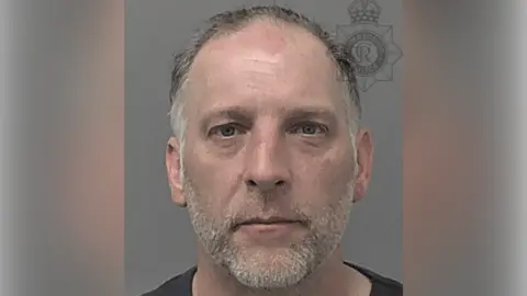 Humberside Police A police custody photo of Nigel Bull. He has short grey hair, a receding hairline and grey facial stubble. He wearing a black t-shirt and is looking at the camera. The wall behind him is grey.