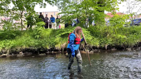 A woman wearing waders and a red lifejacket, and carring a net, walks through a river while a small gathering of people watch from the bank