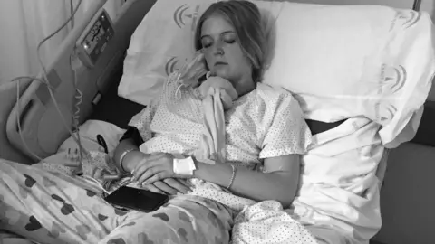 Lauren Jean A black and white photo of Lauren Jean lying in a hospital bed in a hospital gown while sleeping. 