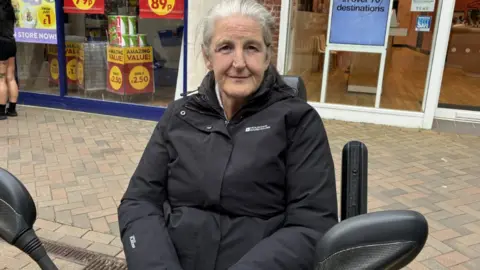 An older lady with grey hair sat in a black mobility scooter. She's wearing a black mountain warehouse coat. She's sat on a high street with shops behind her.