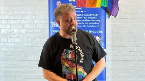 Mark Nicholas A man stands in front of a mic with a large blue sign behind him. A rainbow pride flag is hung over one side of the sign. The man is mid laugh.