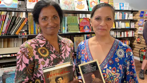Juliet Parkin/BBC Setta and Lazy Sicarmann were photographed with their books at the Oxfam Library in Hove, East Sussex. Seeta bought a biography of Houdini while Lizzzie bought special notes and confessions from the justified sinner. Seeta wears a pink and green pink dress, while Lizzzie wears a blue, red and yellow dress.