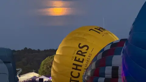 A few hot air balloons filling up with air at night. One is a yellow Thatchers branded on and is mainly in the eyeline. The moon can be seen glowing orange high in the sky.