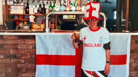 Richard Dowell Richard standing in front of the bar which has a large England flag draped at the front. He has a pint in his hand, a large England hat on, England white t shirt and Union Flag shorts on
