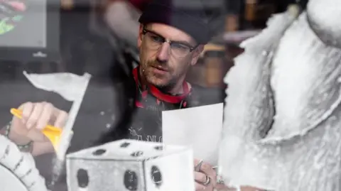 Man wearing glasses and a dark beanie hat behind a window where he is creating a snowscape artwork.