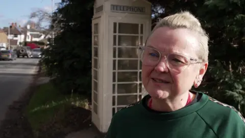 A woman with blond hair, tied in a bun, stands in front of a mid-20th century cream-coloured telephone box by the side of a quiet village street. 