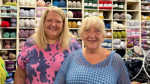 Two women with short blonde hair are standing in front of a shelf filled with different colours of wool.
