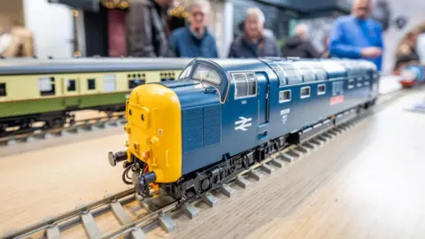 A close up of a yellow and blue diesel locomotive. It is on a section of track laid on a table. Just out of focus is a group of people.