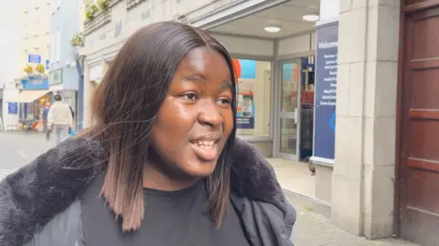 Esther has shoulder length dark brown hair. She is wearing a black t-shirt and a black jacket.