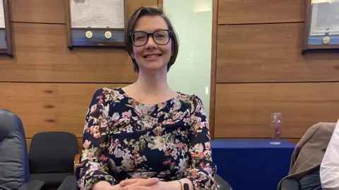Fiona Cole Fiona Cole. She has shirt brown hair, wearing glasses and floral top. Her hands are folded on a desk.