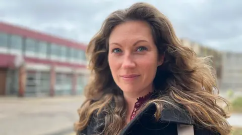 Emma Baugh/BBC A woman with blue eyes stands on the street in front of one of the old nightclubs. She has brown hair past her shoulders and is wearing a black jacket.
