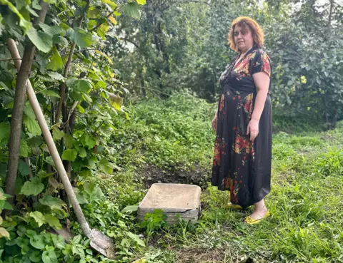 BBC/ Woody Morris Irina's daughter Nino Elizbarashvili stands next to a suitcase and a shovel in the garden
