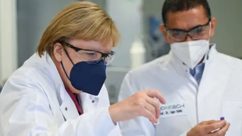 Getty Images Ugur Sahin, founder and CEO of the biotechnology company BioNTech shows German Chancellor Angela Merkel dummy ampoules of the BioNTech/Pfizer COVID-19 vaccine during her visit to the BioNTech Vaccine Production Plant on August 19, 2021 in Marburg, Germany.