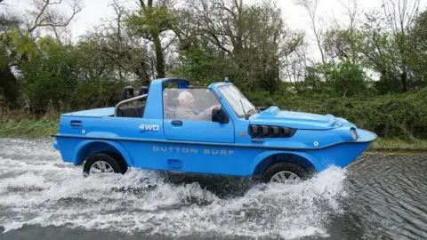 PA Media A blue amphibious car drives through flood water