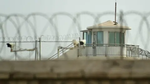 A view over a wall topped with a barbed wire, towards a guard tower, fences and CCTV security cameras