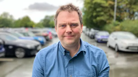 A fair-haired man wearing a blue shirt, standing in front of tree-lined single-storey car park, which is full.