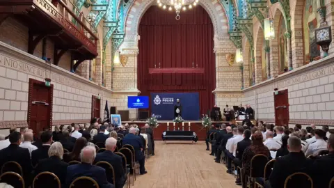 Jenny Kirk/BBC Another view of the civil service as dozens of people sit in rows in a hall, with a high-ceiling, with an aisle down the middle. The people have their backs turned, facing the stage.