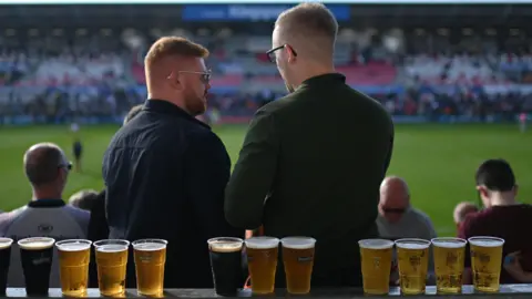 Getty Images Two men at a sporting event stand in front of a row of pints of beer along a rail.