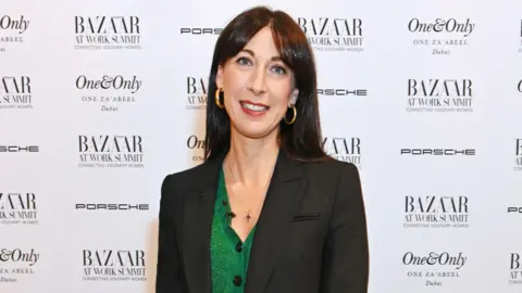 Dave Bennett/Getty Images Samanthan Cameron con un cárdigan verde bajo un blazer negro, en un evento de revista con el tablero promocional detrás de ella. Ella está sonriendo y vistiendo aretes de oro.