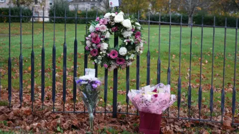 A wreath and two bunches of flowers left against a fence in Bellshill. Behind the fence is a stretch of grass, a hedgerow and a number of trees