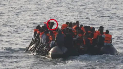 Crown Prosecution Service An image of an overcrowded dinghy at sea. Dozens of figures can be seen, with some wearing orange life vests. There are waves in the water, which is grey, and the people have their backs to the camera.
