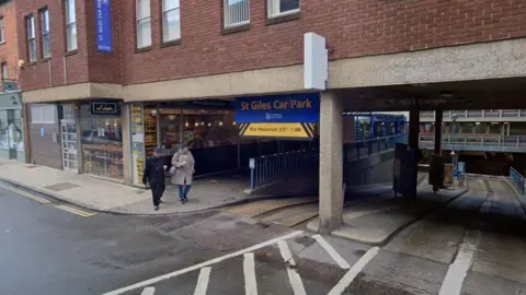 Google Two people are walking past the entrance to St Giles Car Park in Norwich. There is a ramp down to the car park and one coming up to the exit. There are shops on the street next to it.