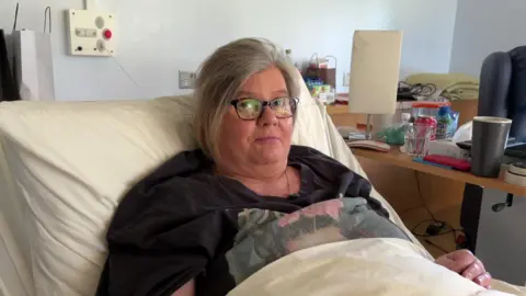 A woman lying in a hospice bed. there are some cups and a kettle on the bed beside her. She is wearing glasses.