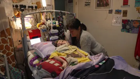 A woman stands over her a man's bed. He is hooked up to ventilators and machines.