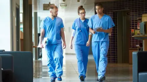 Three junior doctors walking down a hospital corridor while wearing blue scrubs.