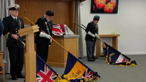 Three men in military uniforms carrying standards have lowered them to the ground at a funeral. There is a coffin on a plinth which is draped with a Union Jack flag and there is a photo of the man as a young sailor in a frame.