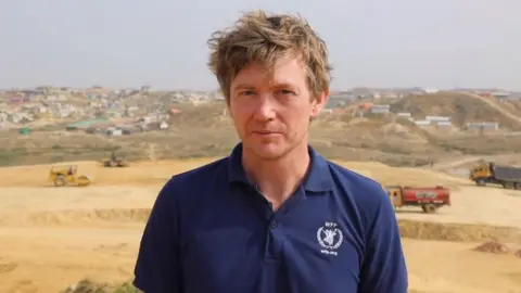 A man with brown hair looking at the camera, he is wearing a navy polo top