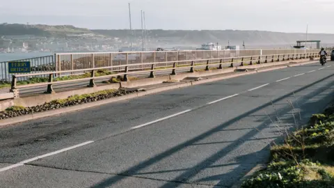 A grey road with white lines and grey fencing with green bushes either side and a number of boats in the background. 