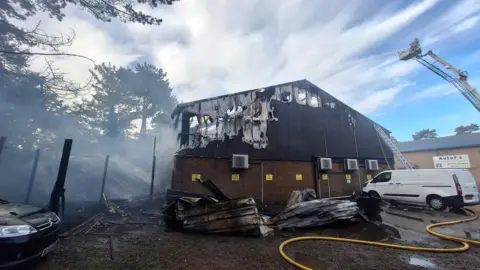 Hertfordshire Fire and Rescue Service Large parts of the side of the building are damaged. There are holes in the wall and the skeleton of the building is visible. An aerial platform shooting a jet of water is to the right. 