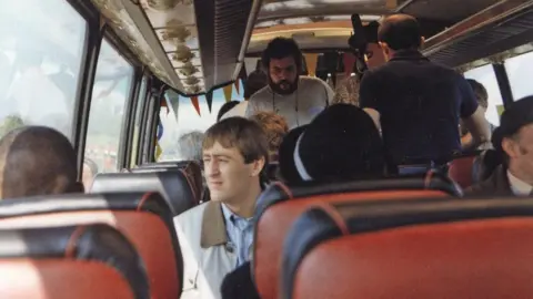 Interior of a coach with rows of red seats, including Rodney Trotter seated by the window among other passengers during a journey.