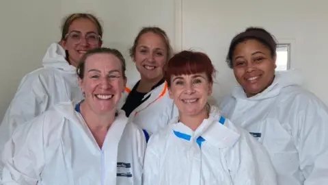 Victoria Gordon Five women are smiling at a camera. They are all wearing white hazmat suits, and are standing in a white room.