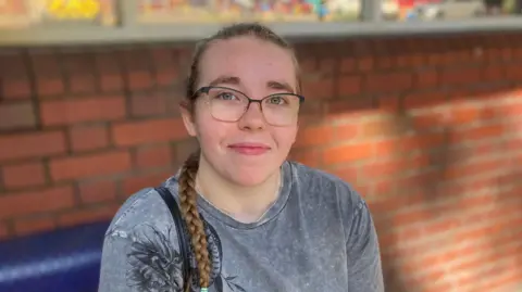 Alex Pope/BBC Megan Hughes has long hair in a plait over one shoulder. She wears glasses and is sitting on a blue bench in front of a brick wall.