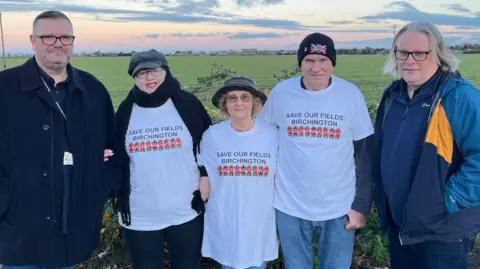 Three people in white shirts that say "save our fields Birchington" on them. There are also two men other side of the three in navy coats.